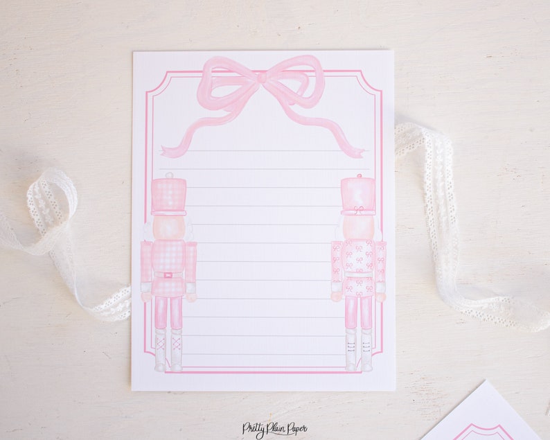 K&ouml;nnte beinhalten: Rosa und wei&szlig;es bedruckbares Briefpapier mit zwei Nussknacker-Soldaten und einer rosa Schleife oben. Das Papier hat liniertes Schreibfeld und den Text "Pretty Plain Paper" unten.