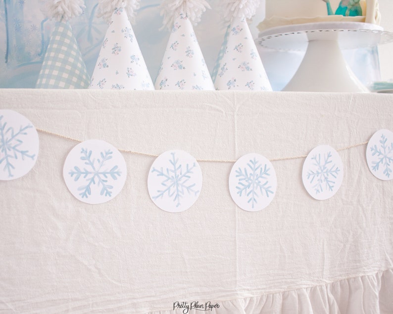 Pu&ograve; includere: Un tovaglia bianca con una ghirlanda di fiocchi di neve blu appesa ad essa. La ghirlanda &egrave; fatta di cerchi di carta con fiocchi di neve blu dipinti su di essi. La ghirlanda &egrave; infilata con lo spago.