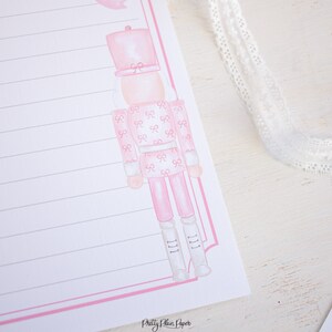 K&ouml;nnte beinhalten: Rosa und wei&szlig; bedrucktes Briefpapier mit einer rosa Aquarell-Illustration eines Nussknackers. Das Briefpapier hat einen rosa Rand und liniertes Papier. Der Text "Pretty Plain Paper" befindet sich am unteren Rand der Seite.