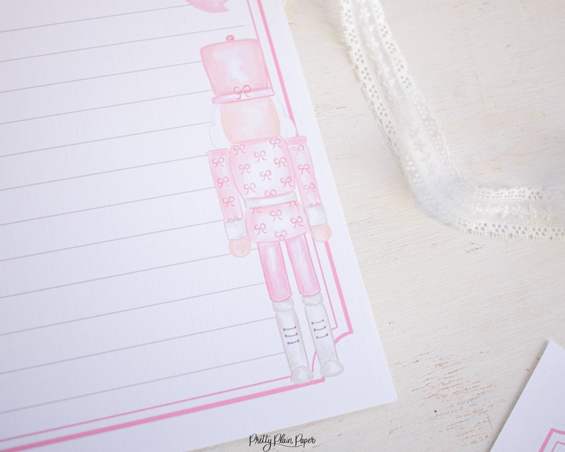 K&ouml;nnte beinhalten: Rosa und wei&szlig; bedrucktes Briefpapier mit einer rosa Aquarell-Illustration eines Nussknackers. Das Briefpapier hat einen rosa Rand und liniertes Papier. Der Text "Pretty Plain Paper" befindet sich am unteren Rand der Seite.