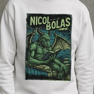 Peut inclure: Sweat-shirt blanc avec un motif d'un démon vert lisant un livre, avec le texte "NICOL BOLAS" en vert. L'illustration est de style vintage, bande dessinée, avec un fond sombre et texturé.