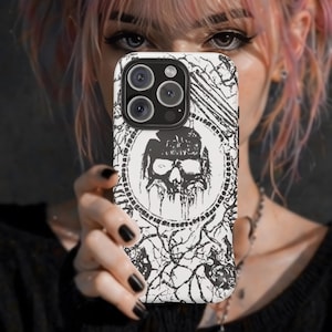 May include: A white and black phone case featuring a skull design within a circular frame. The case has a textured pattern and is held up in front of a person's face. The phone case has a black camera module.