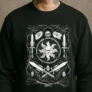 May include: Black sweatshirt with a white graphic. The design includes a sun face, crossed swords, daggers, a skull, and other symbols. The artwork is framed by branches and decorative elements. The design is in white.
