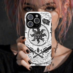 May include: A white phone case with a black sun design, crossed swords, and other graphic elements. The case has a black camera module and is held up in front of a person's face.