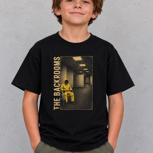 May include: A black short-sleeve t-shirt featuring a rectangular graphic print. The design depicts a figure in a yellow hazmat suit sitting in a dimly lit, beige-walled corridor with the text THE BACKROOMS printed vertically in yellow.