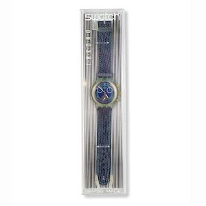 Könnte beinhalten: Eine Swatch Chrono-Uhr in der Originalverpackung. Die Uhr hat ein blaues Zifferblatt mit goldfarbenen Akzenten und ein blaues strukturiertes Lederarmband. Die Uhr befindet sich in einer transparenten Kunststoffbox mit dem Swatch-Logo und dem Text "Swiss Made".