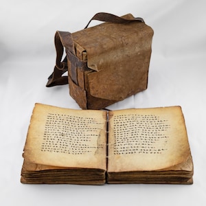 Puede incluir: Un libro antiguo abierto con texto manuscrito y páginas envejecidas se encuentra debajo de un bolso de cuero marrón desgastado con una correa para el hombro. Las páginas del libro están amarillentas y el bolso de cuero muestra signos de desgaste.