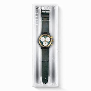 Può includere: Un orologio Swatch in una custodia di plastica trasparente. L'orologio ha un cinturino in pelle verde scuro e una lunetta color oro. Il quadrante dell'orologio è verde con tre sotto-quadranti bianchi. La custodia ha la scritta "Swatch".