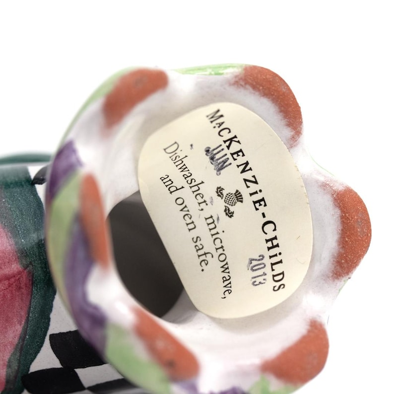 May include: A colorful ceramic object with a floral design. The rim features a pattern of red, green, and purple. A label reads "MacKenzie-Childs" and "Dishwasher, microwave, and oven safe."