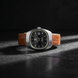 Könnte beinhalten: Eine silberfarbene Tissot Seastar Automatik-Uhr mit schwarzem Zifferblatt und braunem Lederarmband. Die Uhr hat ein rechteckiges Zifferblatt mit Datumsanzeige, vor dunklem Hintergrund.