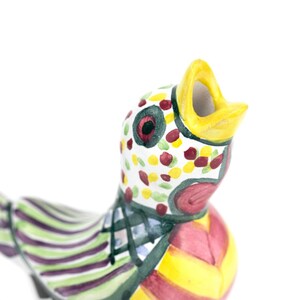 May include: A colorful ceramic bird figurine with an open beak. The bird features a white head with red, yellow, and green spots. The body is striped with green, purple, and yellow. The beak is yellow.