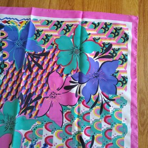 May include: A square scarf with a vibrant floral pattern. The scarf features large flowers in shades of blue, pink, and teal, set against a geometric background with a pink border. The design includes various shapes and colours.