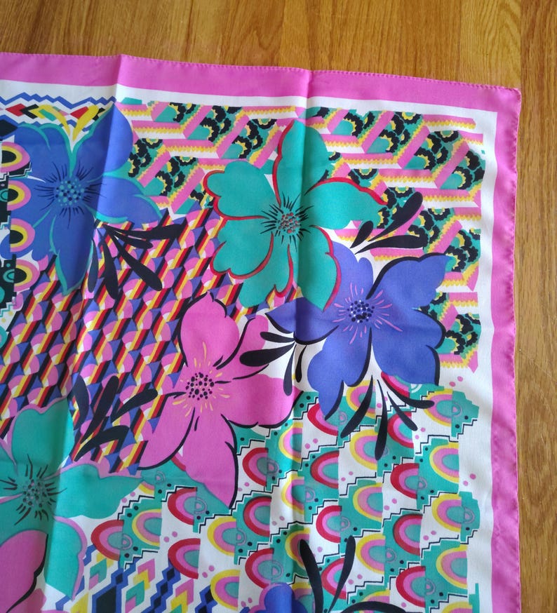 May include: A square scarf with a vibrant floral pattern. The scarf features large flowers in shades of blue, pink, and teal, set against a geometric background with a pink border. The design includes various shapes and colours.
