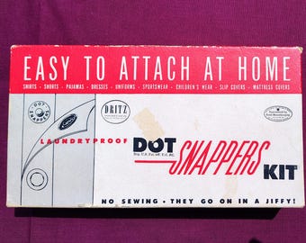 May include: A white box with red lettering that reads "Easy to Attach at Home" and "Dot Snappers Kit". The box features a laundry-proof design and instructions for attaching the snaps. The box is for a kit of laundry-proof snaps.