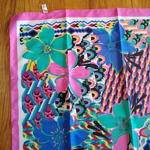 May include: A patterned square scarf with a bold floral and geometric design. The scarf showcases large flowers in teal, pink, and blue, set against a white background with a pink border. Geometric shapes in various colours add to the design.