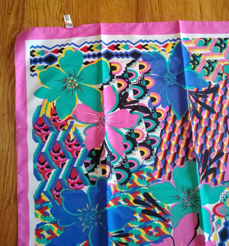 May include: A patterned square scarf with a bold floral and geometric design. The scarf showcases large flowers in teal, pink, and blue, set against a white background with a pink border. Geometric shapes in various colours add to the design.