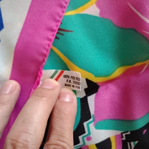 May include: A vibrant floral fabric with a pink border. The fabric has a tag that reads "100% POLYES R.N. 76593 MADE IN ITAL". The design features teal, pink, yellow, and black shapes.