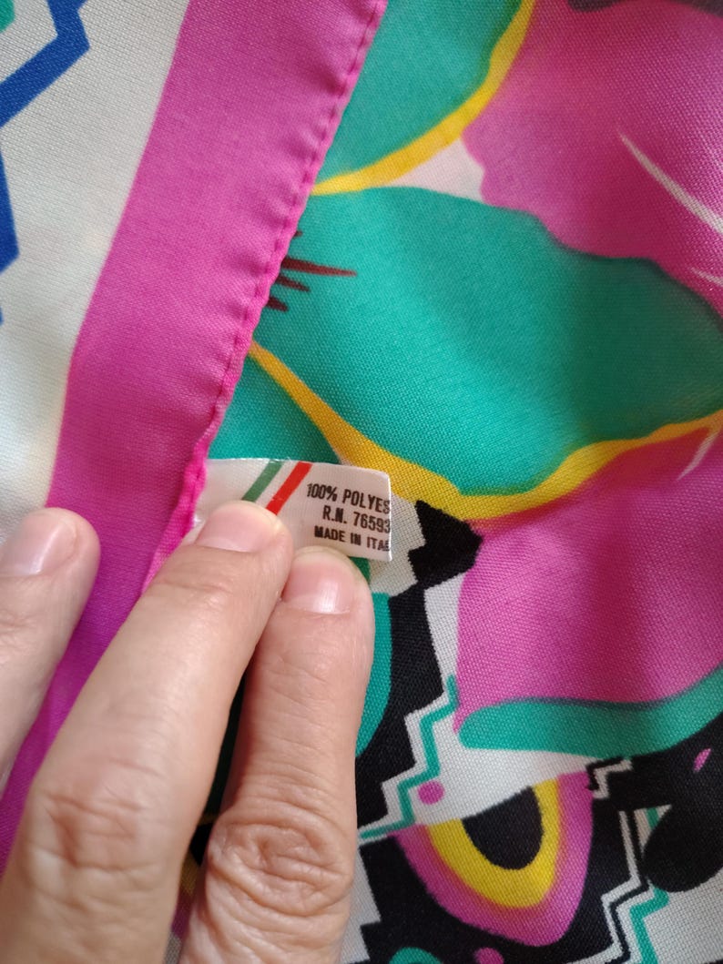 May include: A vibrant floral fabric with a pink border. The fabric has a tag that reads "100% POLYES R.N. 76593 MADE IN ITAL". The design features teal, pink, yellow, and black shapes.
