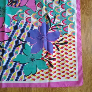May include: A vibrant silk scarf with a floral design. The scarf showcases large flowers in pink, purple, and teal, set against a geometric background with a pink border. The scarf is made of a lightweight material.