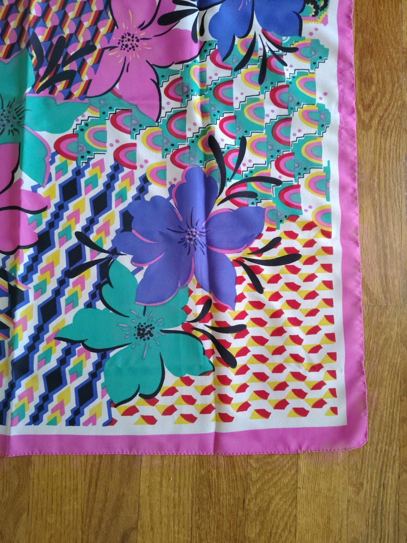 May include: A vibrant silk scarf with a floral design. The scarf showcases large flowers in pink, purple, and teal, set against a geometric background with a pink border. The scarf is made of a lightweight material.