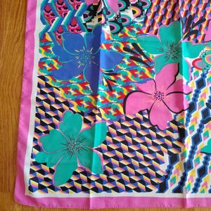 May include: A patterned silk scarf with a bold floral and geometric design. The scarf showcases large flowers in blue, pink, and teal, set against a backdrop of geometric shapes in various colours. A pink border frames the design.