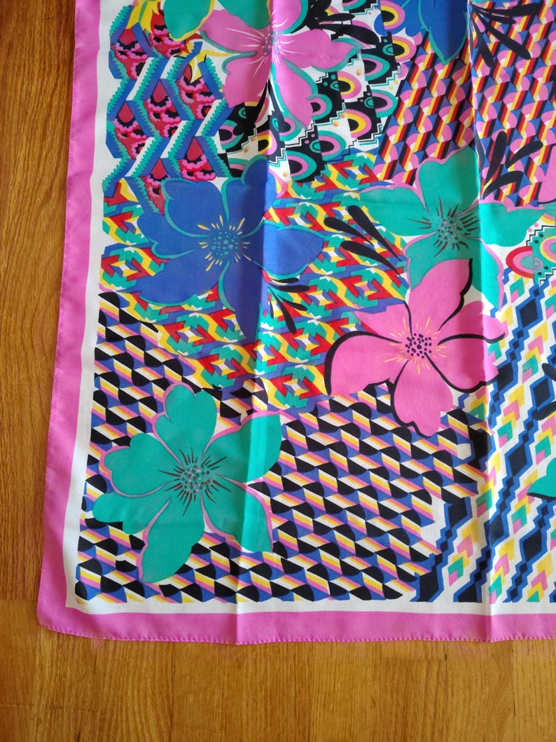 May include: A patterned silk scarf with a bold floral and geometric design. The scarf showcases large flowers in blue, pink, and teal, set against a backdrop of geometric shapes in various colours. A pink border frames the design.