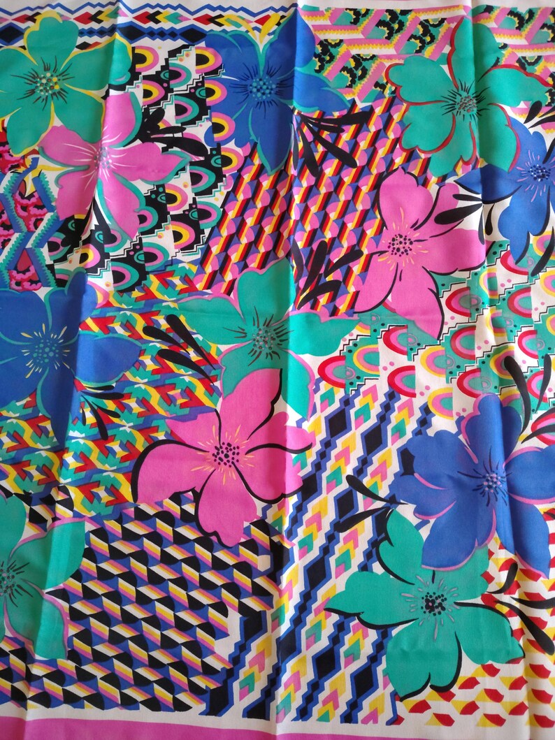 May include: A colourful floral fabric with large flowers in pink, blue, and teal. The design is set against a patchwork of geometric and abstract patterns. The fabric has a white background with a pink border.