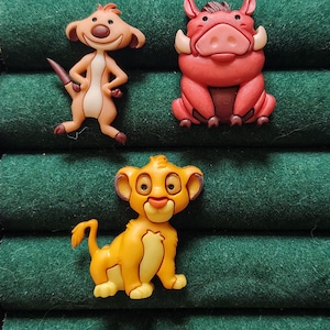May include: Three colorful character figurines from the animated film "The Lion King" are displayed against a green background. The figurines include Timon, Pumbaa, and Simba, each with distinct features and vibrant colors. The figures are made of a hard plastic material.