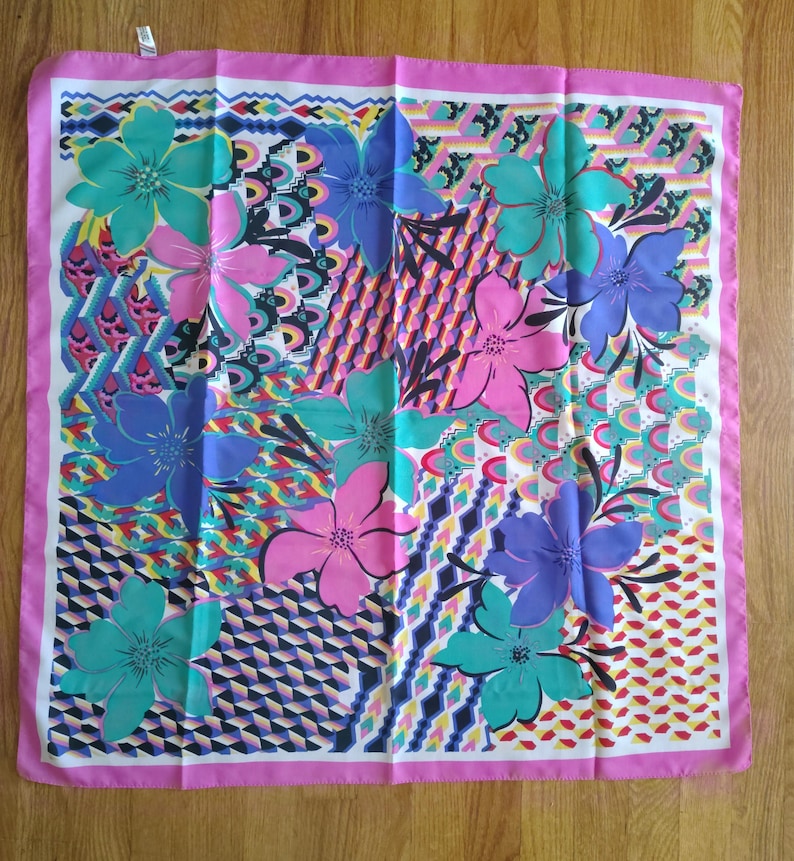 May include: A square silk scarf with a colourful floral design. The scarf displays large flowers in pink, teal, and purple, set against a geometric background. A pink border frames the design. The scarf is likely used as a fashion accessory.