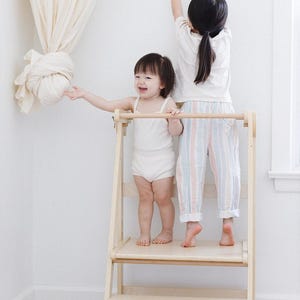 May include: Two young children are shown in a bright, minimalist setting. One child stands on a wooden learning tower, while the other reaches for a decorative element. The scene is bathed in natural light, creating a warm and inviting atmosphere.