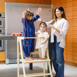 May include: A light wood step stool with two children and an adult. One child is wearing a blue jumpsuit, the other is in white. The adult is wearing a white shirt and jeans. They are in a kitchen with wood cabinets and stainless steel appliances.