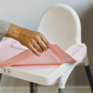 May include: A pink high chair mat being placed on a white high chair. The mat is made of a waterproof material and is designed to protect the chair from spills and messes.