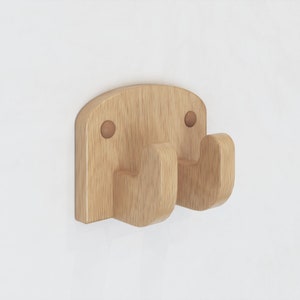 May include: A wooden wall hook with a light brown finish. The hook has a rectangular back with rounded top corners and two curved hooks. Two small, round, brown pegs are visible on the back. The hook is designed for hanging items.