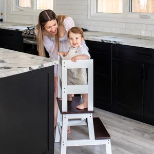 May include: A white wooden learning tower with a brown wooden step. A young child is standing on the top step, looking at the camera. An adult woman is standing behind the child, smiling.