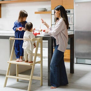 May include: A wooden step stool with a safety rail, with two children standing on it. A woman and a red apple slicer are also in the kitchen. The children are wearing blue and white outfits.