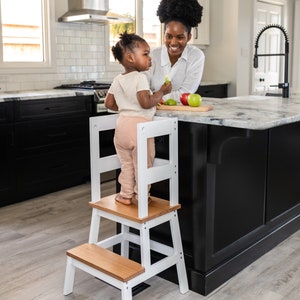 May include: A white wooden learning tower with two steps, a child standing on the top step, and an adult standing behind the child. The child is holding a green apple and the adult is holding a red apple.