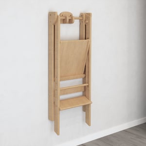 May include: A light wood folding chair mounted on a white wall. The chair is folded up and has a back and a seat.