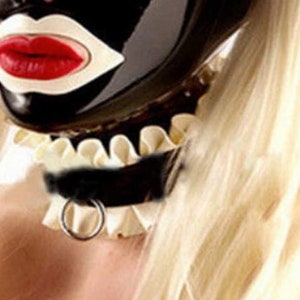 Puede incluir: Una m&aacute;scara de l&aacute;tex negra con un dise&ntilde;o blanco de boca abierta y labios rojos. La m&aacute;scara la lleva una persona con el pelo rubio y un collar blanco con volantes con una gargantilla de cuero negro y un anillo de plata.