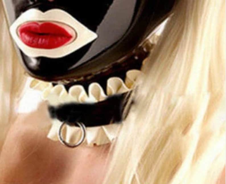 Puede incluir: Una m&aacute;scara de l&aacute;tex negra con un dise&ntilde;o blanco de boca abierta y labios rojos. La m&aacute;scara la lleva una persona con el pelo rubio y un collar blanco con volantes con una gargantilla de cuero negro y un anillo de plata.