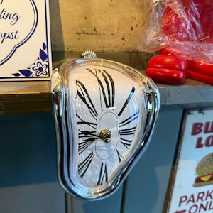 Può includere: Un orologio da tasca argentato e sciolto con quadrante bianco e numeri romani neri. L'orologio ha uno stelo e lancette color oro. L'orologio è un design surrealista, che ricorda un famoso dipinto.