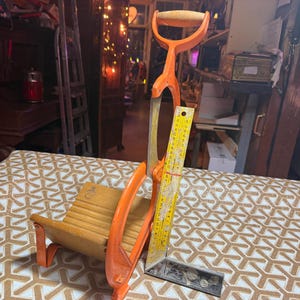 May include: An orange and wood-toned antique woodworking tool set, including a saw, a miter box, and a yellow ruler. The saw has a wooden handle and a metal blade. The miter box is made of wood and has orange accents.