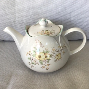 Peut inclure: Une théière en céramique de couleur crème avec un design à bords festonnés et un couvercle assorti. Le corps et le couvercle présentent un motif floral délicat avec des fleurs jaunes et roses, des feuilles vertes et des tiges brunes.