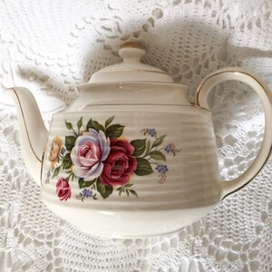 May include: Cream ceramic teapot with a lid and handle, decorated with a floral pattern of red, pink, and yellow roses. The teapot has gold trim and a ribbed texture. A classic tea set item.