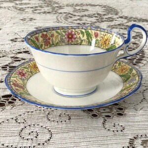 May include: A porcelain teacup and saucer set featuring a cream-coloured base with a decorative border of colourful flowers in shades of pink, yellow, and green. The rim and handle are accented with a thin blue line, and the handle has a curved shape.
