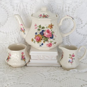 May include: Cream-colored vintage tea set with a teapot, creamer, and sugar bowl. The set features a floral design with pink roses, blue flowers, and green leaves. The teapot has a diamond pattern and a lid. The creamer and sugar bowl also have floral accents.