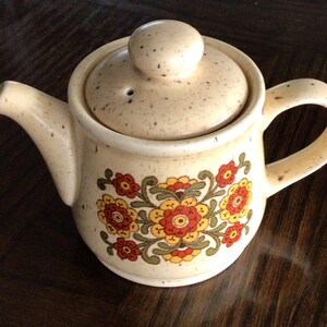 May include: A beige ceramic teapot with a floral pattern in shades of red, yellow, and green. The teapot has a lid with a small knob on top.
