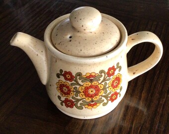May include: A beige ceramic teapot with a floral pattern in shades of red, yellow, and green. The teapot has a lid with a small knob on top.