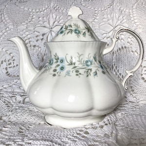 Peut inclure: Une théière blanche avec un motif floral composé de fleurs bleues et vertes. La théière a un couvercle avec un bouton sur le dessus et une poignée sur le côté.