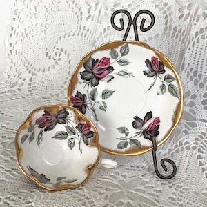 May include: A white teacup and saucer with a black and pink rose pattern and gold trim. The saucer has a gold rim and the teacup has a gold rim and a gold band around the middle.