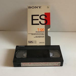 May include: A black Fujifilm VHS video cassette tape positioned in front of its original Sony ES T-120 Dynamicron cardboard sleeve. The sleeve features white, black, and red branding with text indicating 246 metres of tape length.